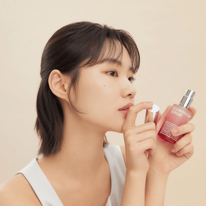 Žena s tmavými vlasmi drží fľašu Blemish Care Serum v jednej ruke a druhou odstraňuje uzáver. Má neutrálny výraz tváre a pozadie je svetlo béžové, čo vytvára jemný a minimalistický dojem.