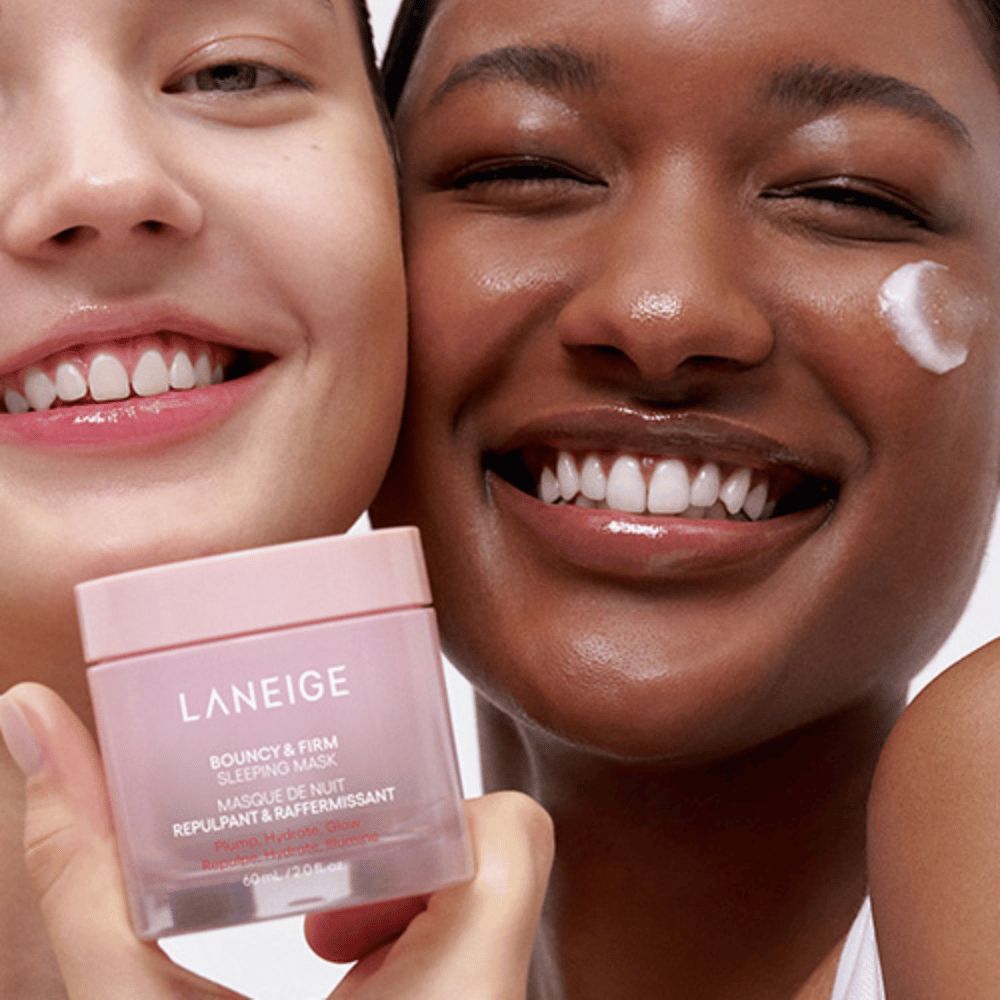 Dvaja ľudia sa usmievajú a držia Laneige Bouncy & Firm Sleeping Mask. Jeden z nich si naniesol masku na líce a produkt je jasne viditeľný v ich rukách. Maska je v ružovej nádobe a je propagovaná ako "Vypnúť, Hydratovať, Žiariť."