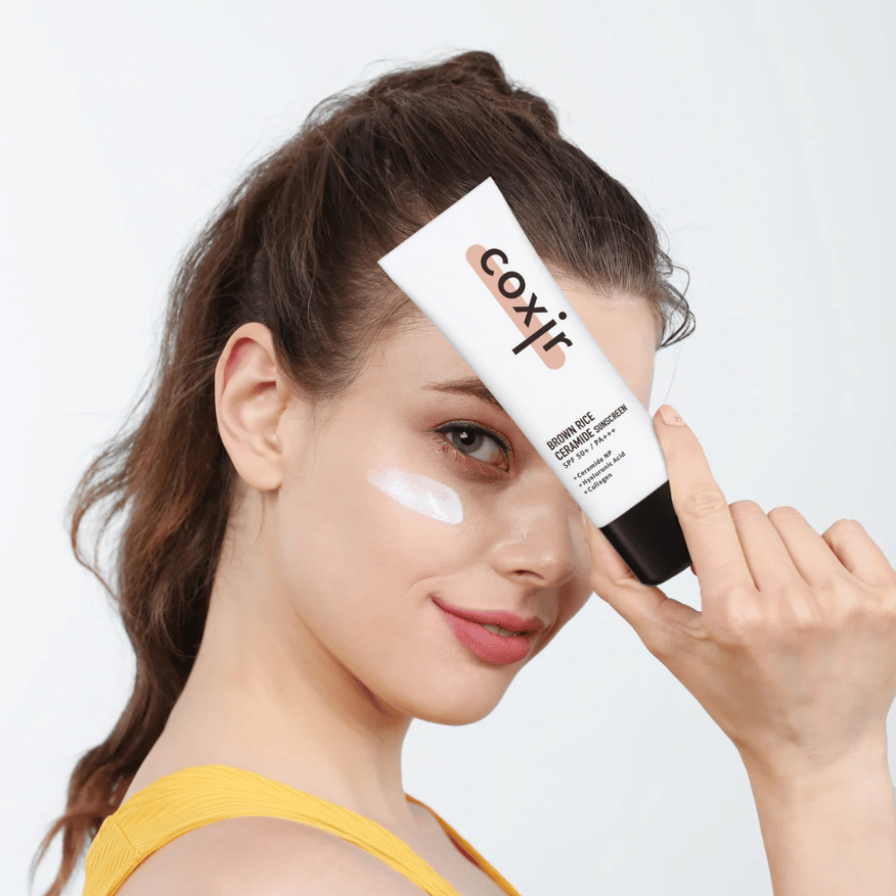 Žena s naneseným opaľovacím krémom na líci drží tubu Coxir Brown Rice Ceramide Sunscreen, SPF 50+ PA+++, pri čele a jemne sa usmieva.