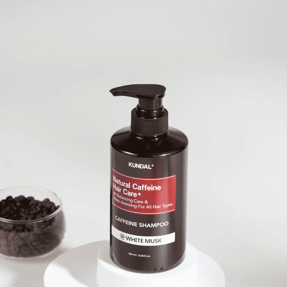 KUNDAL Natural Caffeine Hair Care+ Šampón White Musk v čiernej fľaši s pumpičkou, 500 ml, umiestnený vedľa misky s kávovými zrnami.