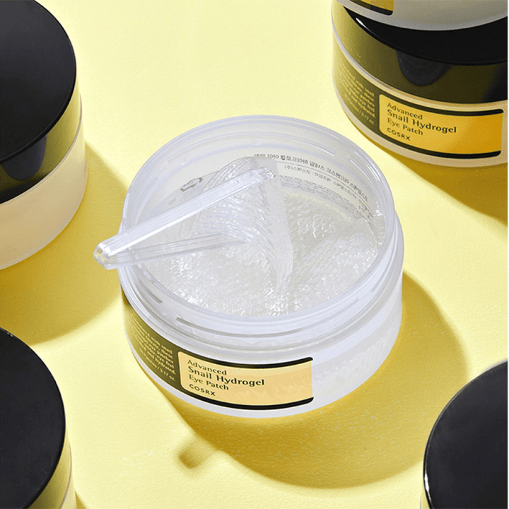 Otvorená nádoba COSRX Advanced Snail Hydrogel Eye Patch s priehľadnými očnými náplasťami a malou špachtľou, obklopená ďalšími uzavretými nádobami na žltom pozadí.