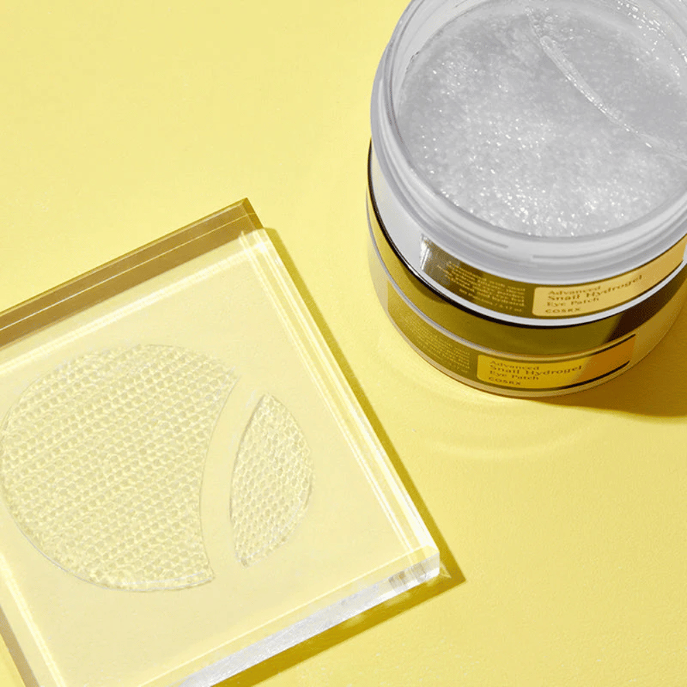 Otvorená nádoba COSRX Advanced Snail Hydrogel Eye Patch stojí na žltom pozadí vedľa priehľadného podložky, kde sú umiestnené dva očné náplasti.