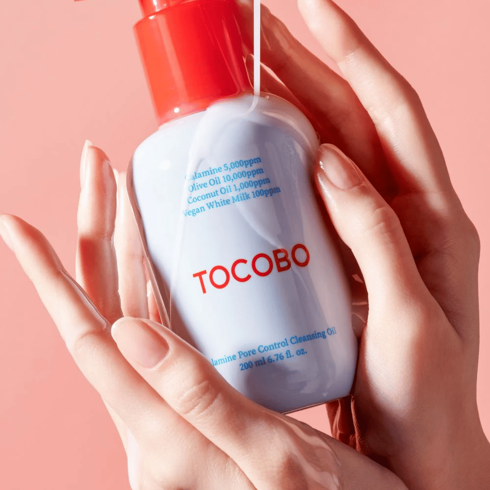 TOCOBO Calamine Pore Control Cleansing Oil držaná v rukách na ružovom pozadí, fľaša s červenou pumpičkou a zoznam ingrediencií sú jasne viditeľné.