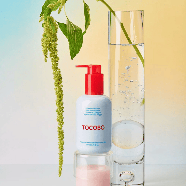 TOCOBO Calamine Pore Control Cleansing Oil s červenou pumpičkou stojí vedľa vysokého skleneného valca naplneného vodou a zelenou rastlinou, na svetlom pastelovom pozadí.