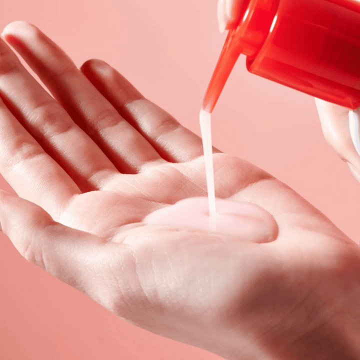 Svetloružový čistiaci olej sa aplikuje z červenej pumpičky do otvorenej ruky na ružovom pozadí.