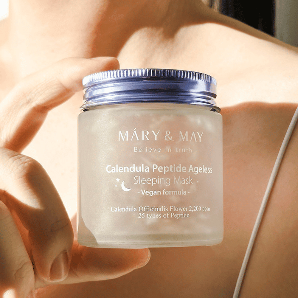 Žena drží pohár s MARY & MAY Calendula Peptide Ageless Sleeping Mask. Pohár je priehľadný s fialovým viečkom a text "Calendula Peptide Ageless Sleeping Mask" je jasne viditeľný. Produkt je vegánska nočná maska s nechtíkom a peptidmi, určená na dodanie mladistvého lesku pokožke počas noci.