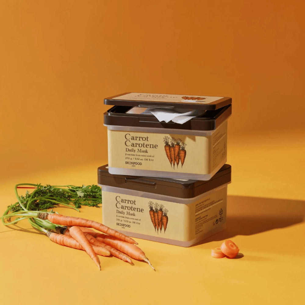 Obrázok zobrazuje tri naskladané balenia "Carrot Carotene Daily Mask" od SKINFOOD, umiestnené na oranžovom pozadí spolu s čerstvými mrkvami. Každá krabica obsahuje pleťové masky obohatené o β-karotén z oleja z mrkvových semien, navrhnuté na každodenné použitie na výživu a starostlivosť o pokožku.