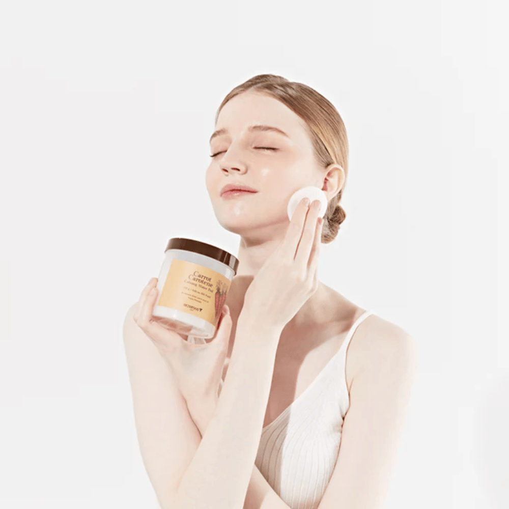 Žena s vyčesanými vlasmi aplikuje kozmetický produkt na svoju tvár pomocou vatového tampónu. Drží veľký pohár označený "Carrot Carotene Calming Water Pad". Jej zatvorené oči a pokojný výraz vyjadrujú pocit relaxácie a potešenia z kozmetickej rutiny.