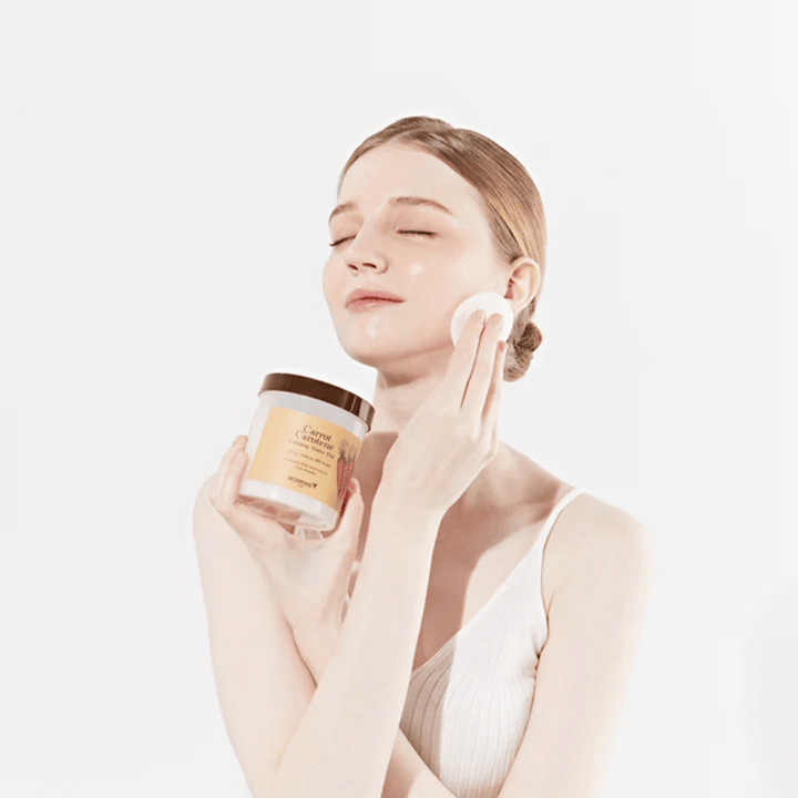 Žena s vyčesanými vlasmi aplikuje kozmetický produkt na svoju tvár pomocou vatového tampónu. Drží veľký pohár označený "Carrot Carotene Calming Water Pad". Jej zatvorené oči a pokojný výraz vyjadrujú pocit relaxácie a potešenia z kozmetickej rutiny.