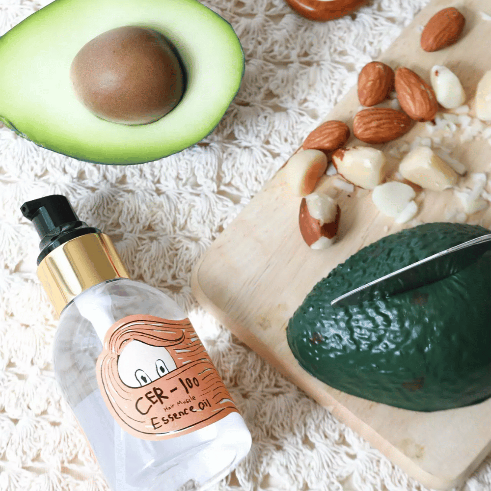 Elizavecca CER-100 Hair Muscle Essence Oil ingrediencie – detailný pohľad s avokádom a rastlinnými olejmi, ktorý ukazuje výživný vlasový olej, ktorý pomáha zmäkčovať a ošetrovať vlasy.