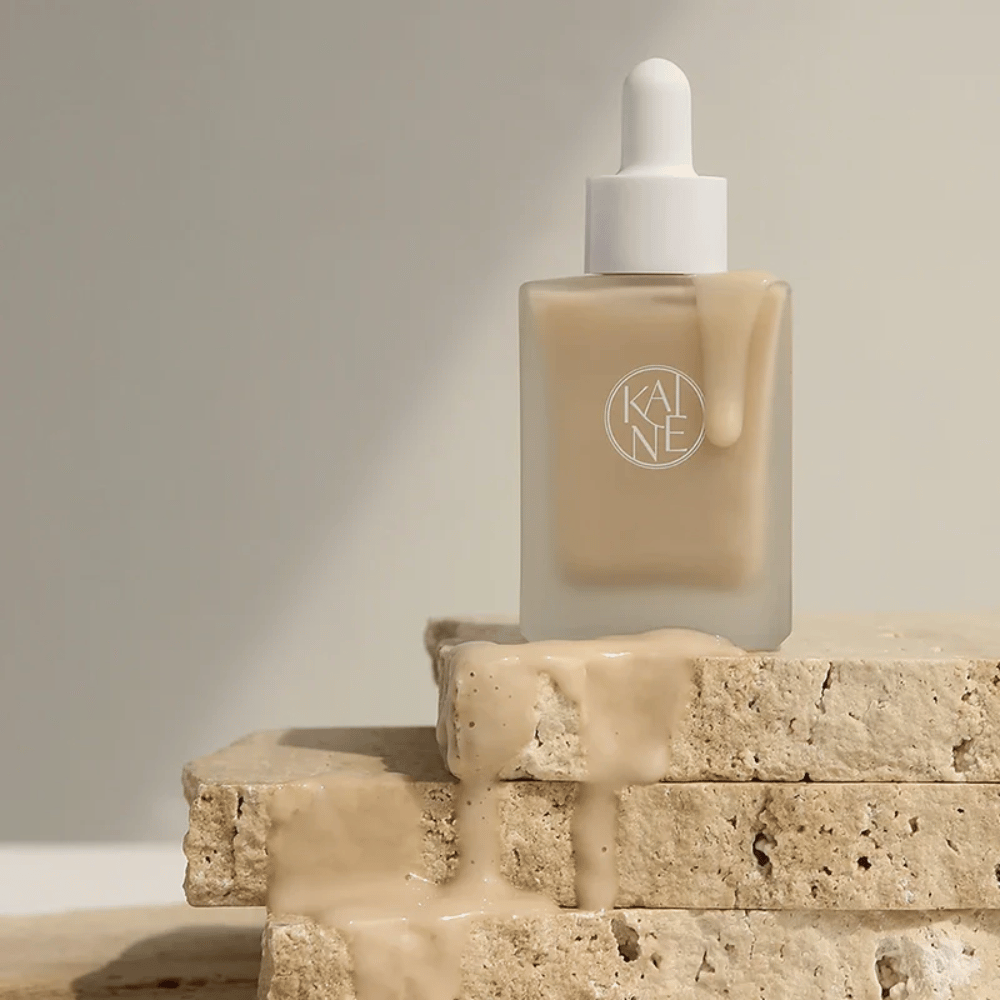 KANE Chaga Collagen Charging Serum v matnej sklenenej fľaši s pipetou. Krémová textúra steká po prírodných kameňoch v mäkkom a minimalistickom svetle.