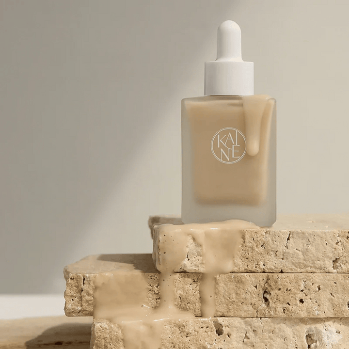 KANE Chaga Collagen Charging Serum v matnej sklenenej fľaši s pipetou. Krémová textúra steká po prírodných kameňoch v mäkkom a minimalistickom svetle.