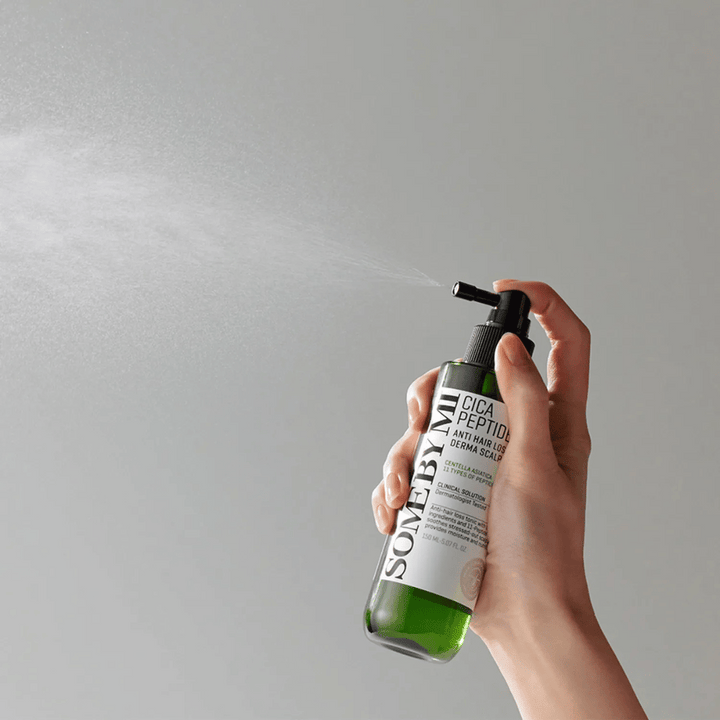 Ruka, ktorá strieka SOME BY MI Cica Peptide Anti Hair Loss Derma Scalp Tonic z zelenej fľaše s čiernou tryskou a bielou etiketou na šedom pozadí.