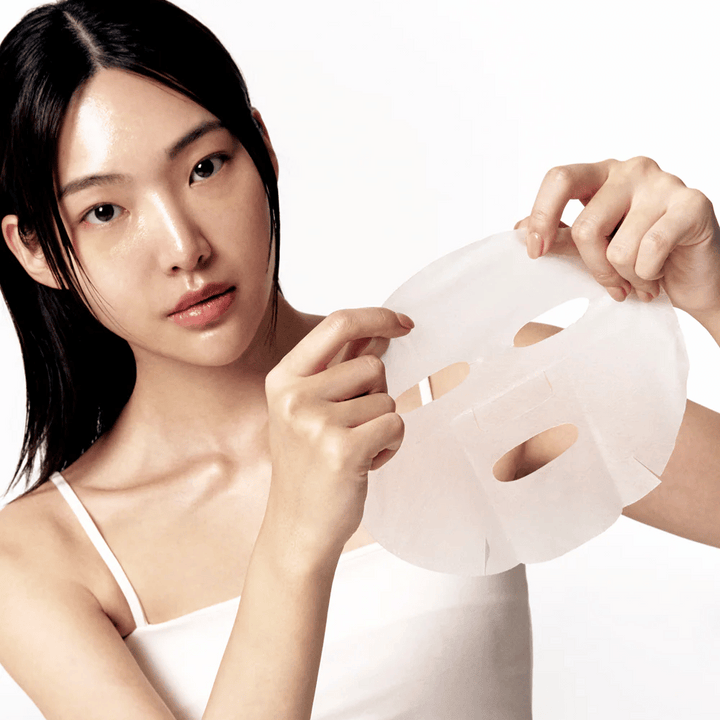 Žena s čistou a hydratovanou pokožkou drží bielu pleťovú masku pred sebou, pripravená ju aplikovať na tvár.