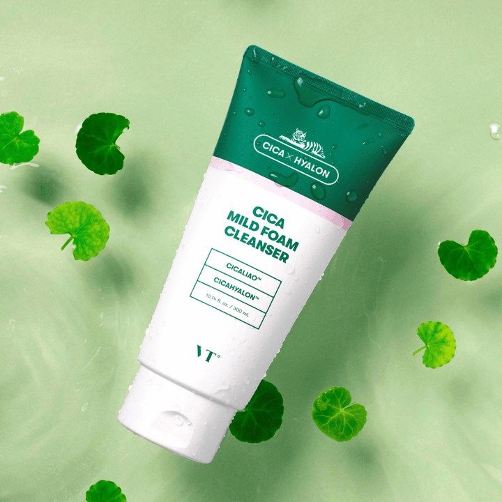 VT Cica Mild Foam Cleanser ležiaca na svetlozelenom podklade s kvapkami vody na balení a obklopená zelenými listami centella asiatica, čo zdôrazňuje upokojujúce vlastnosti produktu.