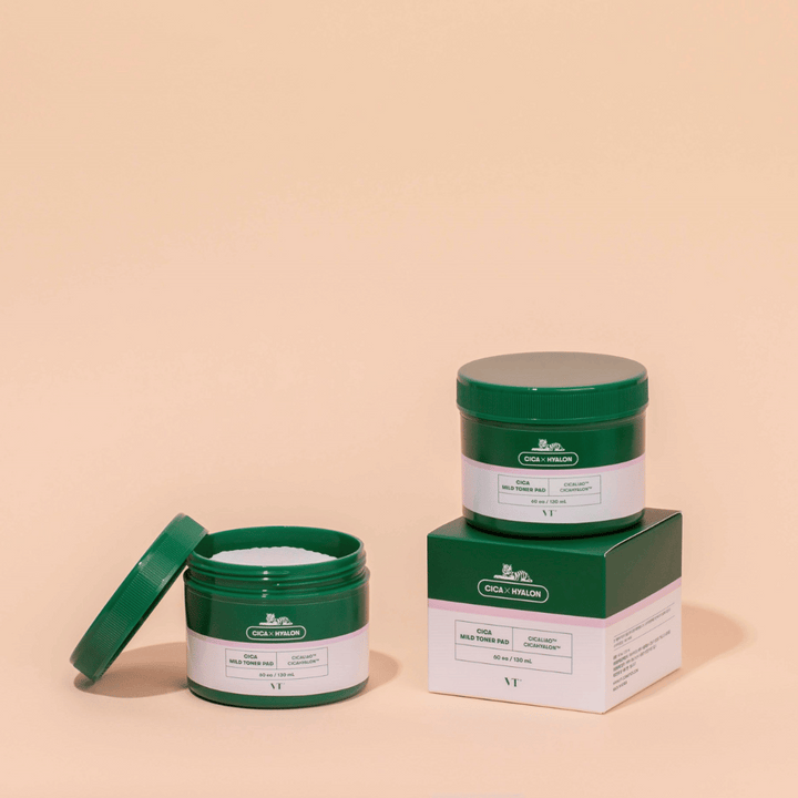 Dve zelené nádoby s VT Cica Mild Toner Pad, jedna otvorená ukazujúca biele tonerové podložky a jedna zatvorená umiestnená na svojej produktovej krabici, na béžovom pozadí.