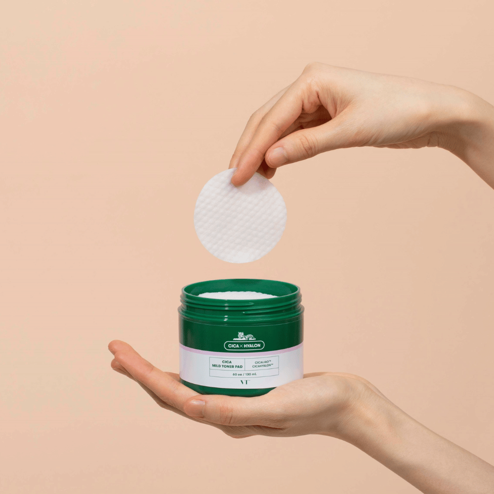 Ruka držiaca zelenú nádobu s VT Cica Mild Toner Pad a dvíhajúca okrúhlu, bielu tonerovú podložku nad nádobou na béžovom pozadí.