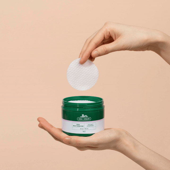 Ruka držiaca zelenú nádobu s VT Cica Mild Toner Pad a dvíhajúca okrúhlu, bielu tonerovú podložku nad nádobou na béžovom pozadí.