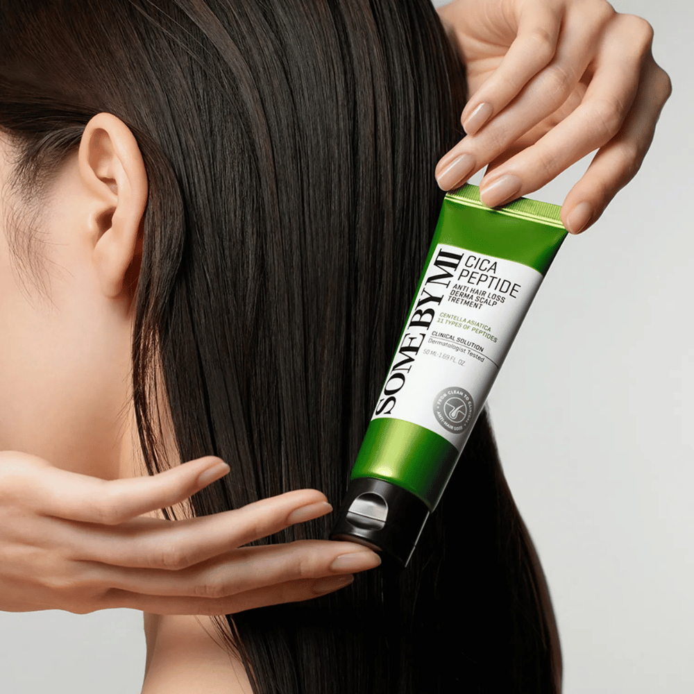 Žena aplikuje SOME BY MI Cica Peptide Anti Hair Loss Derma Scalp Treatment do dlhých, tmavých vlasov so zelenou a bielou tubou.