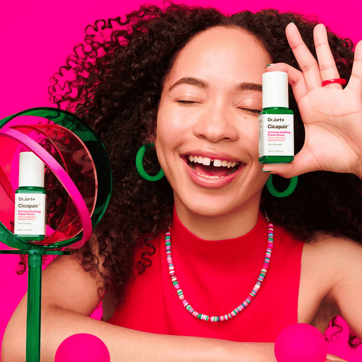 Žena sa široko usmieva, zatiaľ čo drží Dr.Jart+ Cicapair Intensive Soothing Repair Serum pred svojou tvárou. Má na sebe zelené náušnice a červený náhrdelník. Pozadie je svetloružové a zdobené zelenými a ružovými kruhmi.