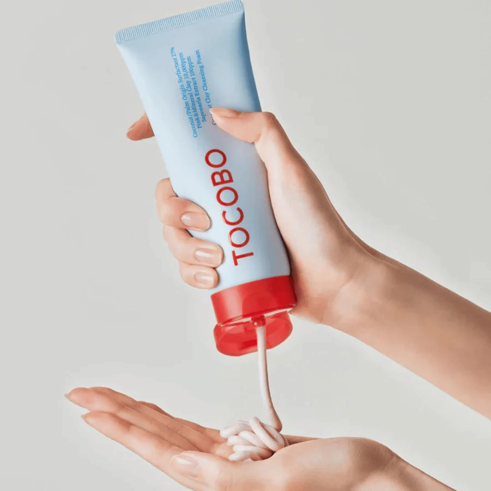 Osoba drží tubu TOCOBO Coconut Clay Cleansing Foam. Tuba je svetlomodrá s červeným uzáverom a textom "TOCOBO" na prednej strane. Produkt sa vytláča z tuby a formuje sa ako šampón v ruke. Pozadie je neutrálne a sústreďuje sa na produkt.