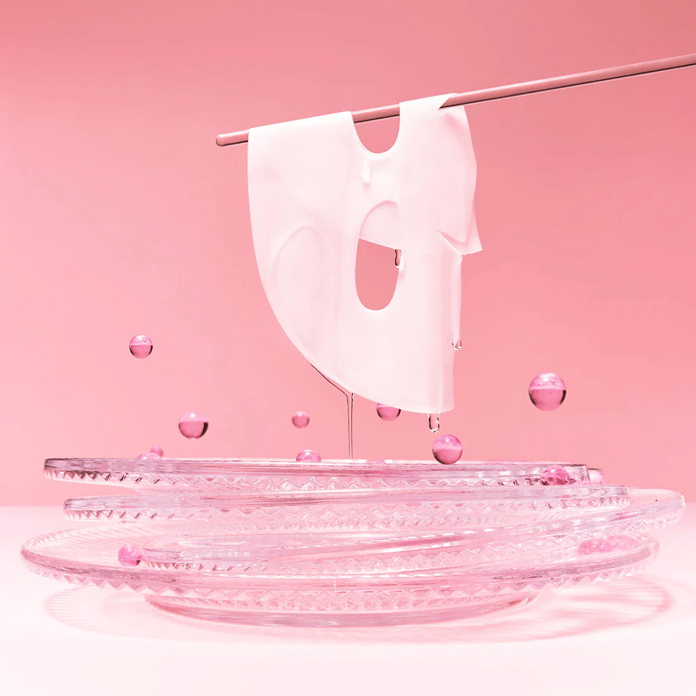 Hydrogélová maska visí na tyči, kvapkajúca sérom. Pozadie je ružové a zrkadlo na spodku odráža ružové bubliny, ktoré obklopujú masku.