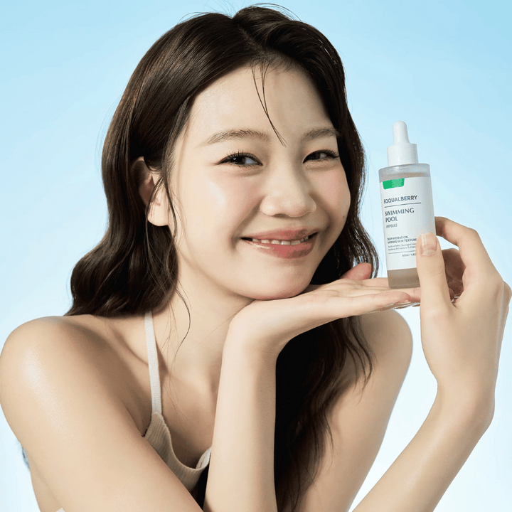 Usmievajúca sa žena drží Eqqualberry Swimming Pool Ampoule, hydratačný produkt starostlivosti o pleť, oboma rukami pred svetlomodrým pozadím.