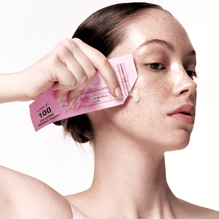 Žena aplikuje krok 1 Collagen Reedle Shot 100 s ružovým jednorazovým balením na líce. Na pokožke je viditeľný číry gél, čo označuje prvý krok dvojkrokovej starostlivosti o pleť.