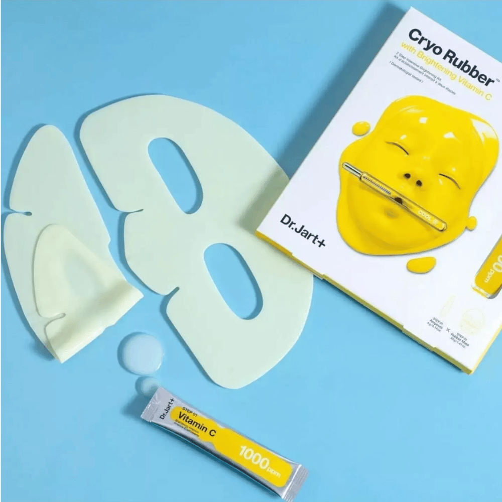 Obrázok kozmetického produktu Dr.Jart+'s 'Cryo Rubber™ s rozjasňujúcim vitamínom C'. Na modrom pozadí je vidieť bielu gumovú pleťovú masku, malú tubu označenú 'Vitamín C 1000ppm' a škatuľu s názvom produktu a obrázkom žltej masky.