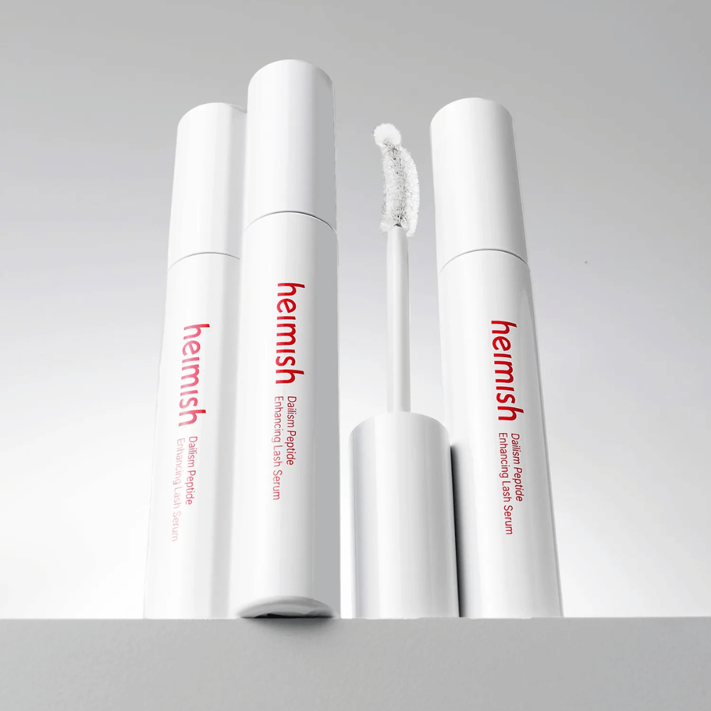 Niekoľko balení Heimish Dailism Peptide Enhancing Lash Serum je zobrazených spolu so zakrivenou aplikačnou kefkou. Obrázok zvýrazňuje elegantný dizajn produktu a presnú kefku na jednoduchú aplikáciu.