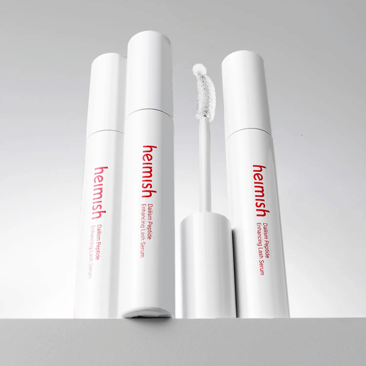 Niekoľko balení Heimish Dailism Peptide Enhancing Lash Serum je zobrazených spolu so zakrivenou aplikačnou kefkou. Obrázok zvýrazňuje elegantný dizajn produktu a presnú kefku na jednoduchú aplikáciu.