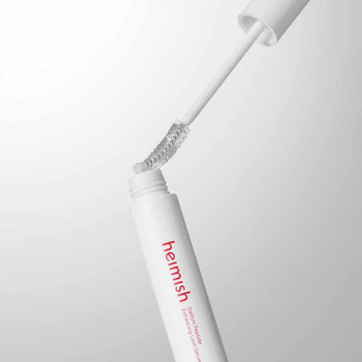 Detailný záber na aplikátor Heimish Dailism Peptide Enhancing Lash Serum. Kefka sa vyťahuje z bielej nádobky a ukazuje transparentné sérum, ktoré sa aplikuje na riasy.