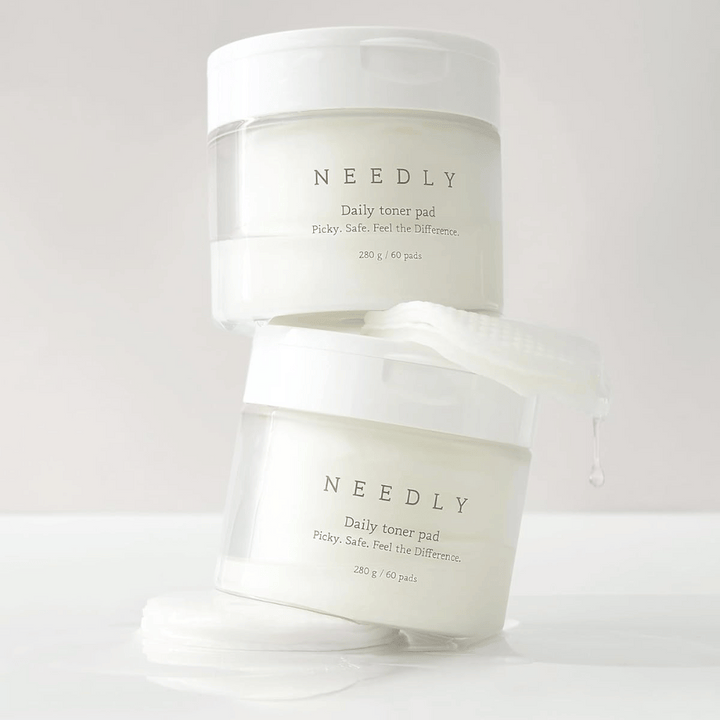 Dve nádoby s Needly Daily Toner Pad naskladané na sebe. Nádoby sú biele s minimalistickým textom a obsahujú 60 tonerových tampónov každá. Jeden tonerový tampón čiastočne vyčnieva z vrchnej nádoby s kvapkajúcou tekutinou.
