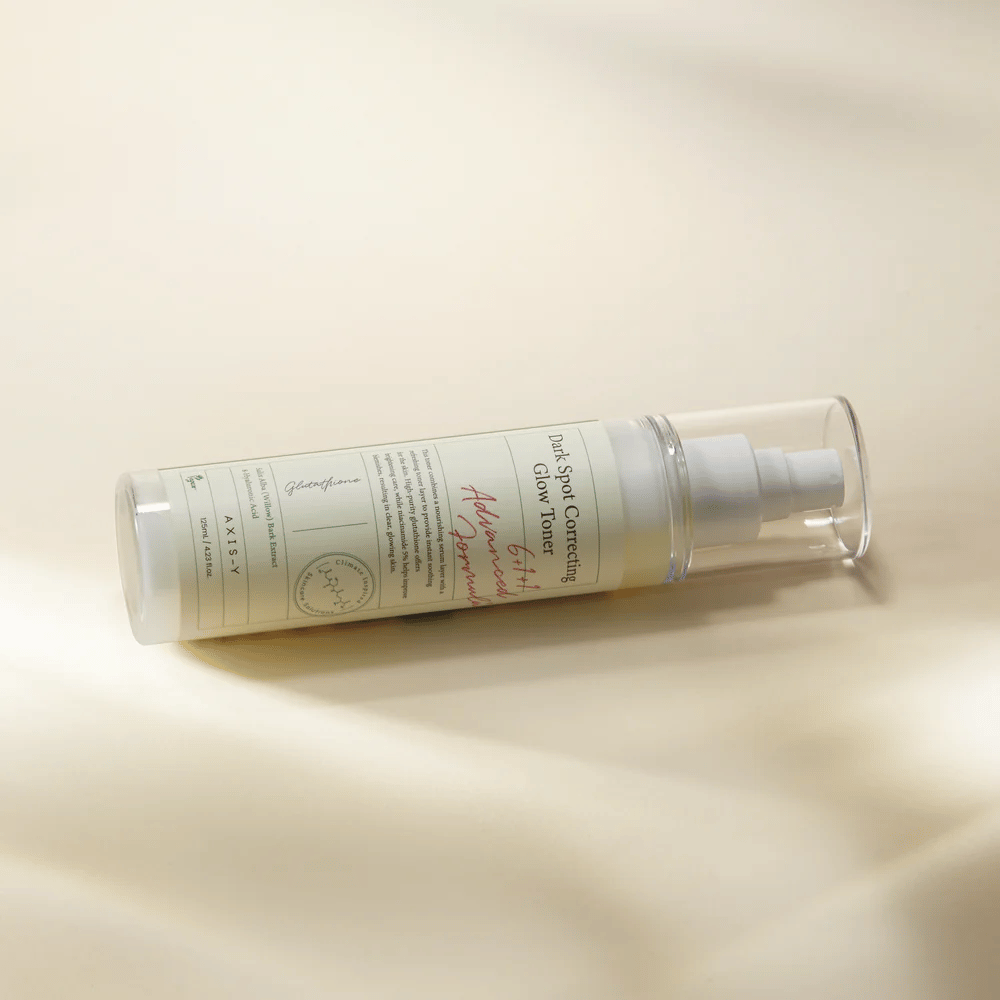 Ležiaca fľaša "AXIS-Y Dark Spot Correcting Glow Toner" umiestnená na svetlom povrchu. Fľaša je priehľadná s bielou etiketou a má bielu sprejovú pumpu. Etiketa zobrazuje informácie o rozjasňujúcich a hydratačných vlastnostiach produktu a obsahuje kľúčové zložky ako Glutathione a Niacinamide.