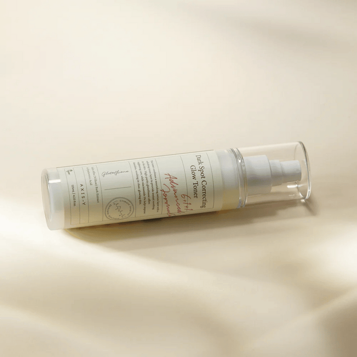 Ležiaca fľaša "AXIS-Y Dark Spot Correcting Glow Toner" umiestnená na svetlom povrchu. Fľaša je priehľadná s bielou etiketou a má bielu sprejovú pumpu. Etiketa zobrazuje informácie o rozjasňujúcich a hydratačných vlastnostiach produktu a obsahuje kľúčové zložky ako Glutathione a Niacinamide.