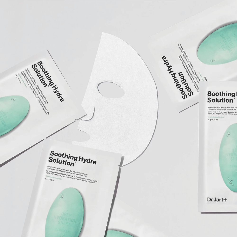 Viacero balení pleťových masiek "Soothing Hydra Solution" od Dr.Jart+ je roztrúsených na svetlom povrchu. Jedna rozvinutá maska je umiestnená v strede, čo ukazuje jej bielu a perforovanú štruktúru.