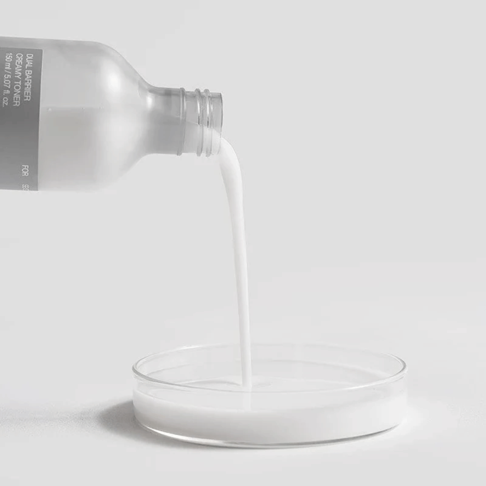 Celimax Dual Barrier Creamy Toner sa opatrne nalieva z fľaše do okrúhleho laboratórneho jamky, s jeho mliečnou a výživnou formulou jasne v popredí.