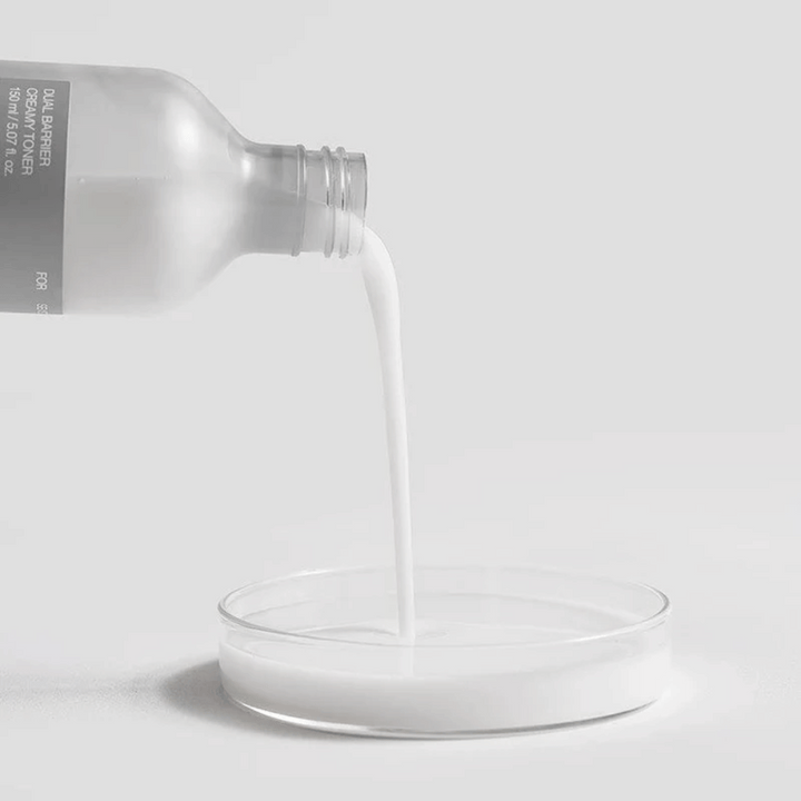 Celimax Dual Barrier Creamy Toner sa opatrne nalieva z fľaše do okrúhleho laboratórneho jamky, s jeho mliečnou a výživnou formulou jasne v popredí.