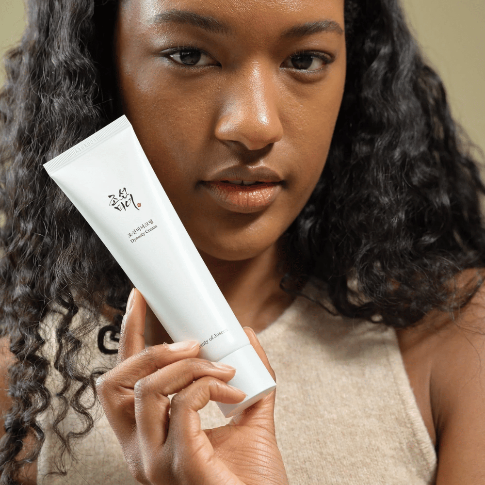 Žena s kučeravými vlasmi drží tubu "Beauty of Joseon Dynasty Cream" blízko svojej tváre, čím zvýrazňuje produkt. Krém, prezentovaný v elegantnej bielej tube, je navrhnutý tak, aby vyživoval a omladzoval pleť, čo zdôrazňuje jeho atraktivitu pre rôzne typy pleti.