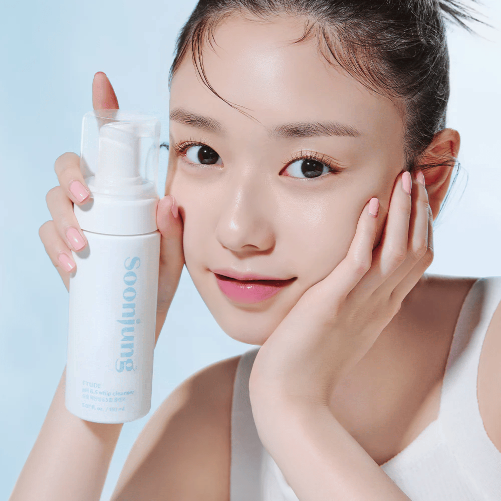 Mladá žena s jasnou pleťou drží bielu pumpovaciu fľašu s textom "Soon Jung pH 6.5 Whip Cleanser" od ETUDE. Usmieva sa na kameru s ľahkým make-upom a sviežim vzhľadom na svetlomodrom pozadí.