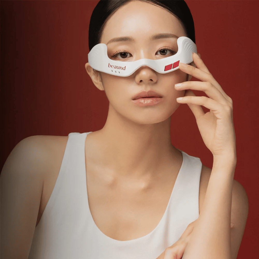Fotografia modelu, na ktorej osoba používa Beaund Eye Rejuv, futuristické LED zariadenie pre oblasť okolo očí, na elegantnom červenom pozadí.