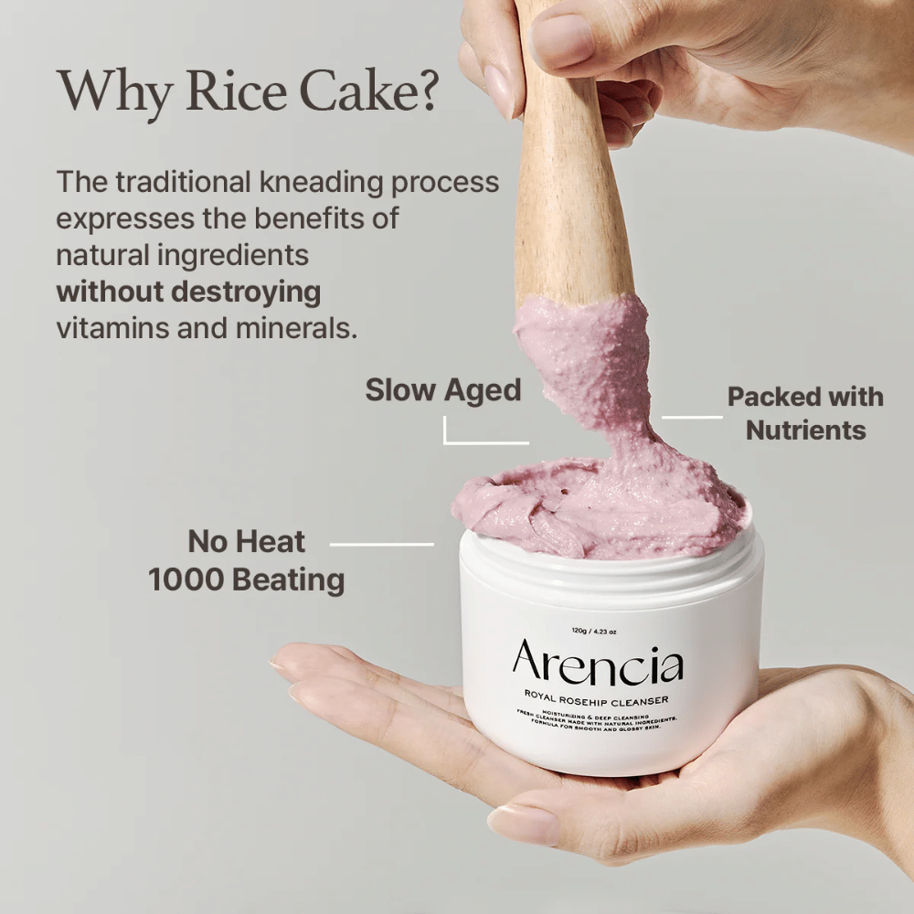 Ruka drží otvorenú nádobu Arencia Royal Rosehip Cleanser, s ružovou, mochi podobnou čistiacou formulou, ktorá je zdvihnutá drevenou špachtľou – ukazuje textúru produktu a tradičnú výrobnú metódu.