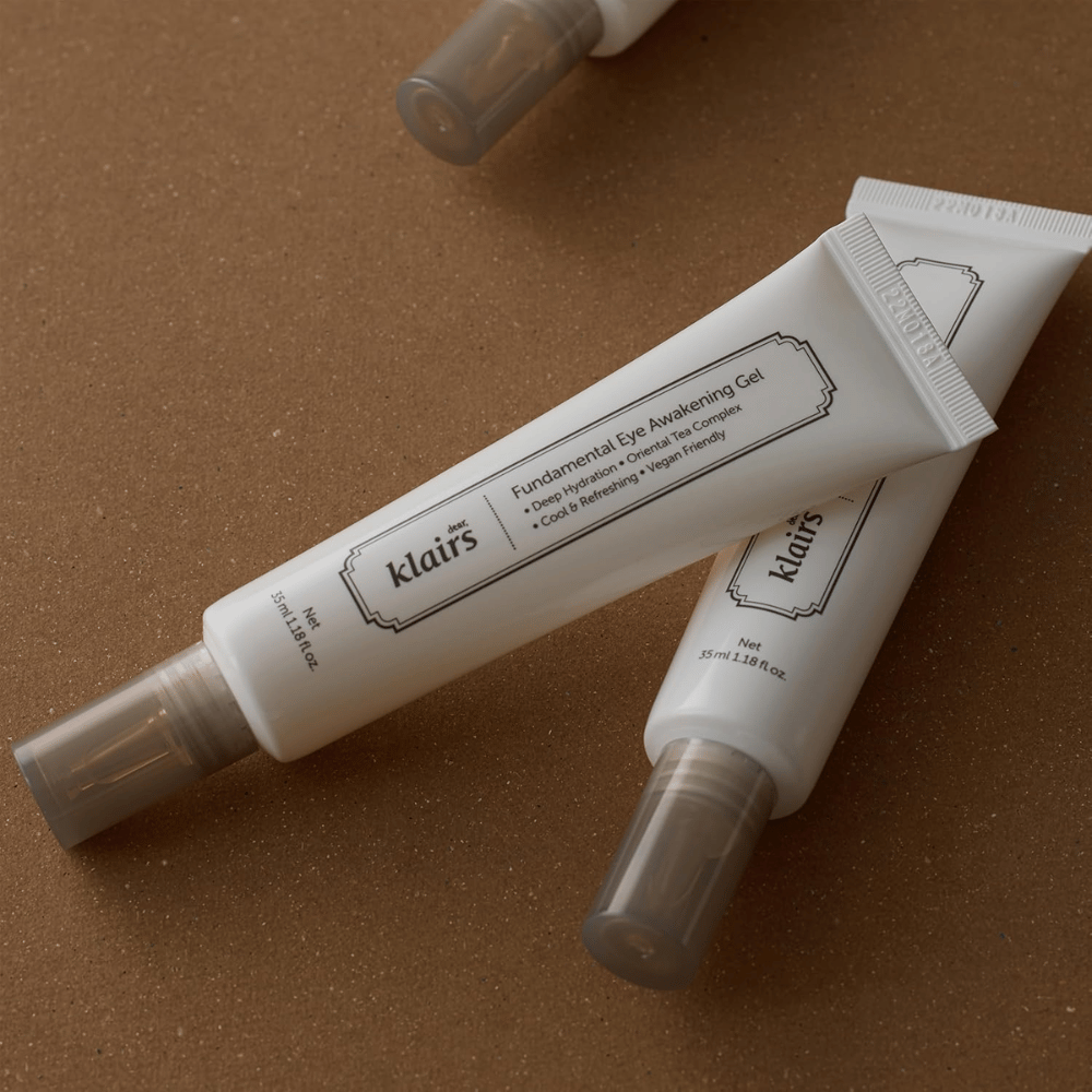 Lifestyle fotografia s dvoma tubami Klairs Fundamental Eye Awakening Gel umiestnenými na teplom hnedom pozadí. Kompozícia zvýrazňuje elegantný dizajn produktu a prémiový dojem, pričom dôraz je kladený na starostlivosť o očné okolie a hydratáciu.