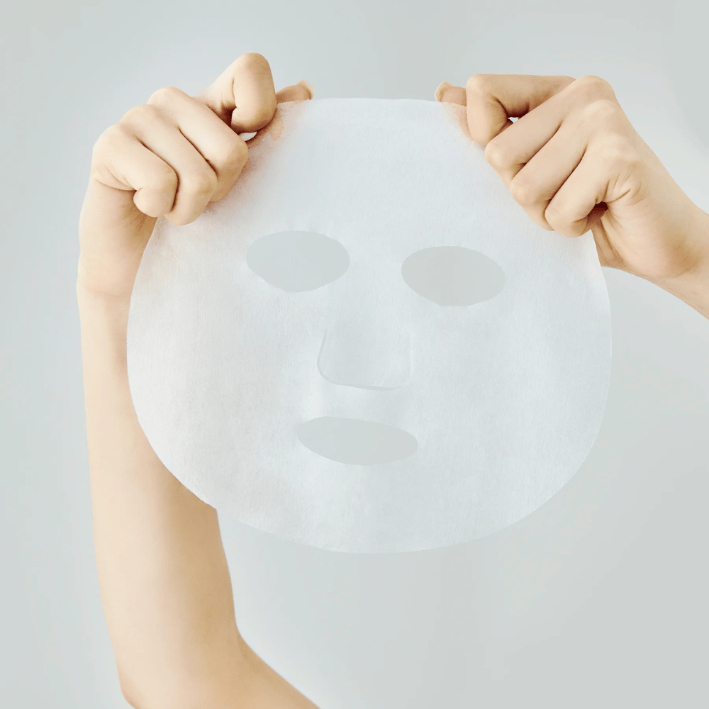Dve ruky držia bielu pleťovú masku na svetlosivom pozadí, s viditeľnými výrezmi pre oči, nos a ústa.