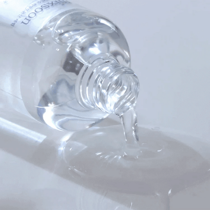 Detailný záber na priehľadné sérum, ktoré sa leje z fľaše Mixsoon Glacier Water Hyaluronic Acid Serum na lesklý povrch.