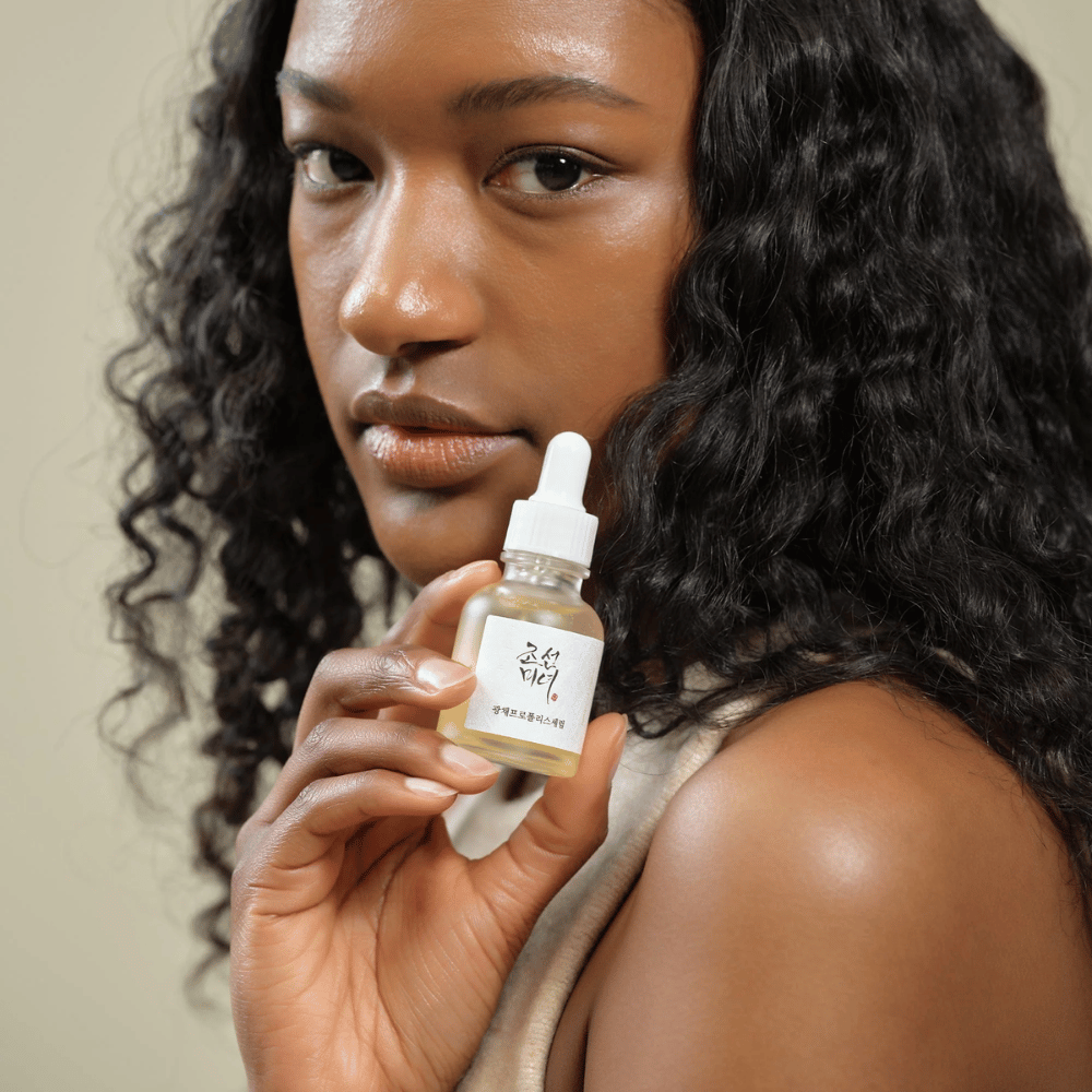 Žena s kučeravými vlasmi drží fľašu Beauty of Joseon Glow Serum s pipetou, hydratačný a rozjasňujúci produkt starostlivosti o pleť.