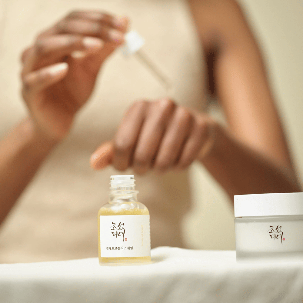 Osoba nanáša Beauty of Joseon Glow Serum s pipetou zo sklenenej fľaše, s nádobou z rovnakej série vedľa na stole.