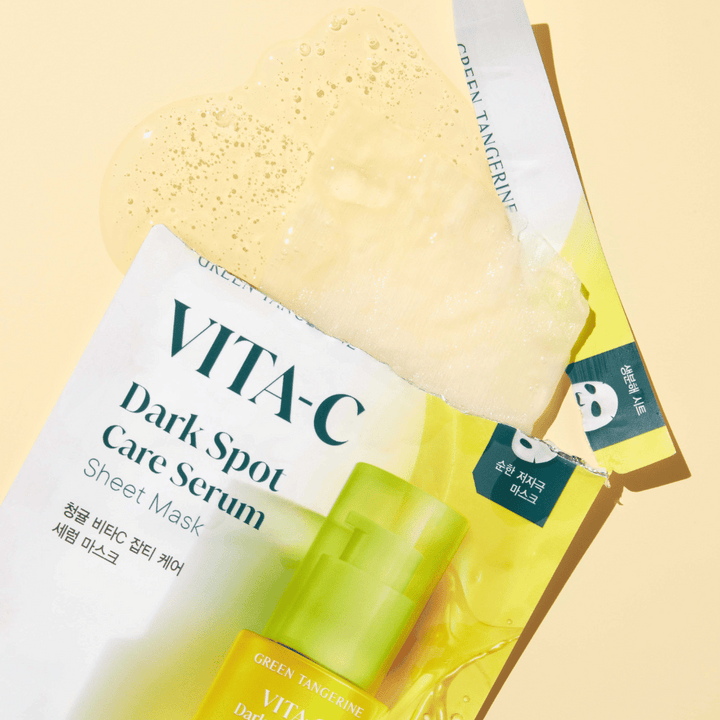 Otvorený obal VITA-C Dark Spot Care Serum Sheet Mask s tvárovou maskou navrchu a zelenou fľašou séra, všetko na žltom pozadí.