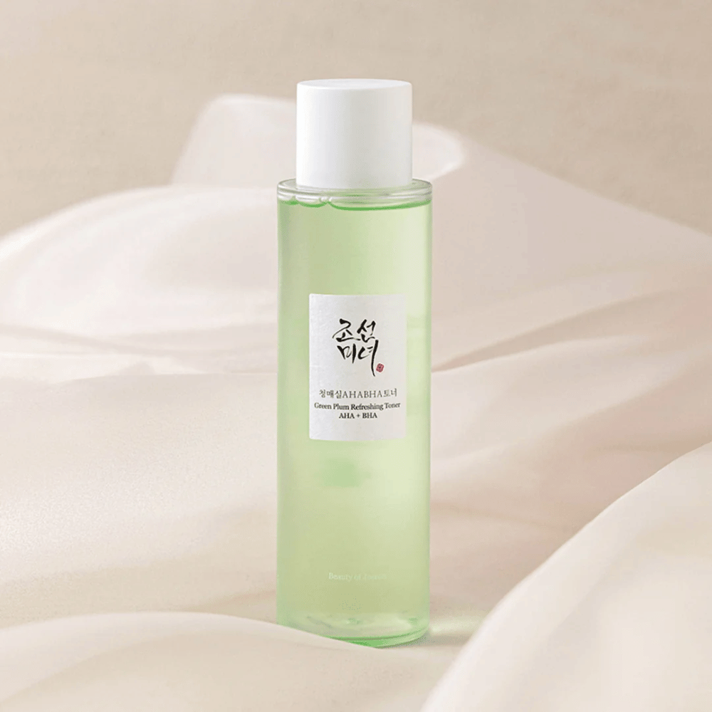 Fľaša s "Green Plum Refreshing Toner" AHA + BHA stojí na mäkkom, svetlom pozadí. Produkt má zelenú tekutinu, ktorá odráža sviežosť a prirodzenosť. Etiketa je biela s čiernymi a červenými prvkami, čo dodáva elegantný a čistý vzhľad.