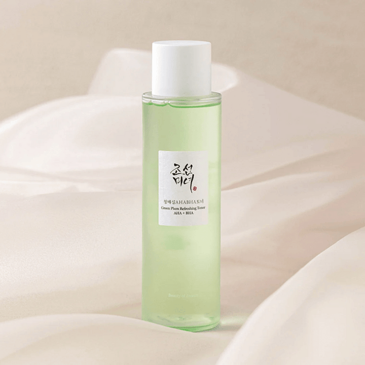Fľaša s "Green Plum Refreshing Toner" AHA + BHA stojí na mäkkom, svetlom pozadí. Produkt má zelenú tekutinu, ktorá odráža sviežosť a prirodzenosť. Etiketa je biela s čiernymi a červenými prvkami, čo dodáva elegantný a čistý vzhľad.